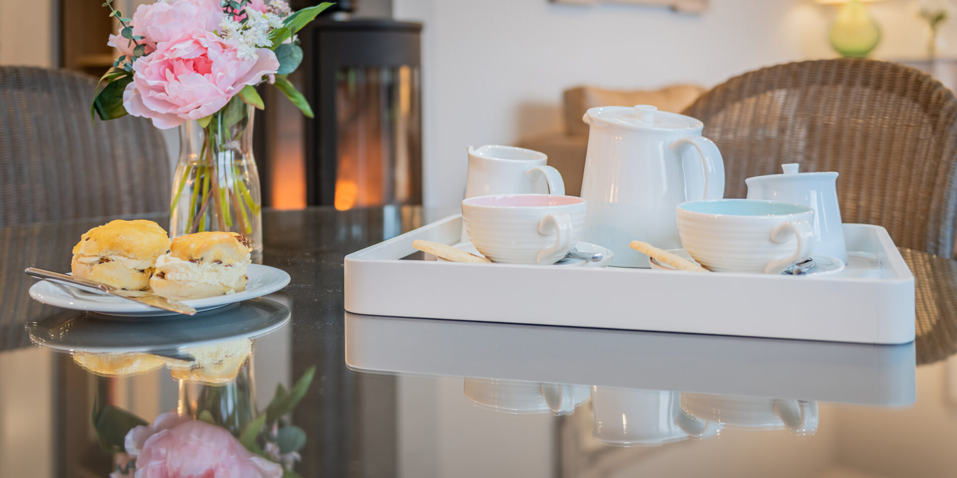 A tea service and a plate of scones rest on top of a glossy black dining table. Next to the scones a vase of pink flowers.